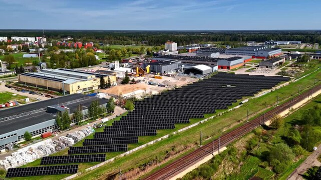 Aerial view of massive solar panel farm beside railway tracks in Jelgava industrial district, Latvia with factories, concrete plant, and city panorama