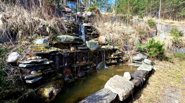Small man made waterfall with tiered stone steps cascading into a backyard pond in early spring
