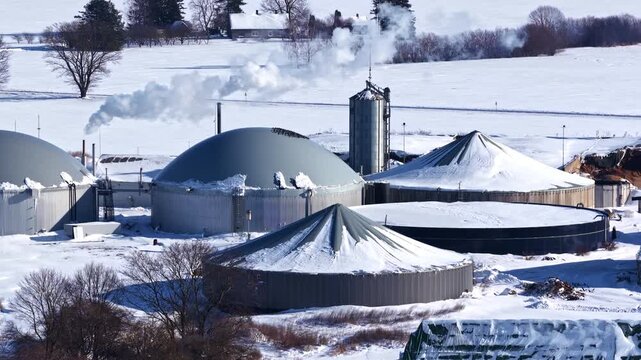 Biogas plant producing renewable energy with smoking chimney in a snowy landscape, aerial view