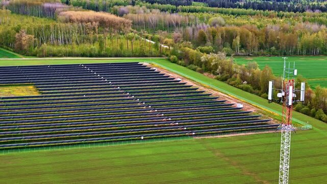 Aerial view of solar panel farm beside telecommunications cell tower surrounded by green agricultural fields and spring forest in Jelgava, Latvia