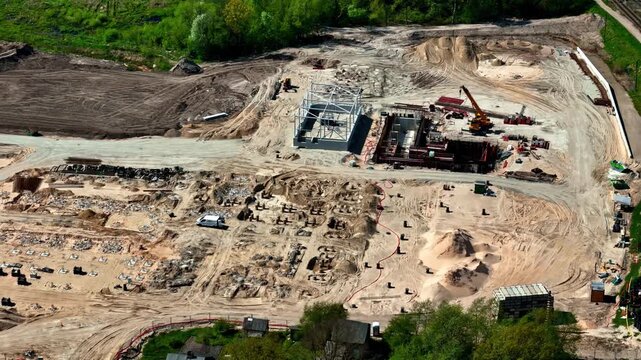 Aerial view of early-stage heavy industrial construction site in Jelgava, Latvia with steel frame structure, orange crane, foundation bolts, and earthworks