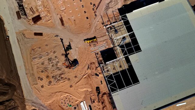 Top-down aerial view of large warehouse construction site in Latvia with foundation pile drilling rig, steel frame structure, and sandy excavated ground
