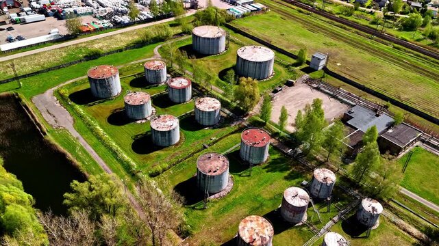 Aerial orbiting view of large Soviet-era cylindrical fuel storage tanks with containment berms beside canal and railway in Latvia