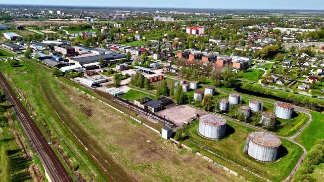 Aerial pullback revealing Soviet-era cylindrical fuel storage tanks beside railway tracks with Jelgava city panorama visible in background, Latvia