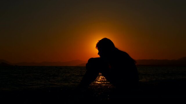 Silhouette of a sad woman sitting alone at sunset. Lonely and sad woman sitting alone on the beach, her silhouette outlined by the sun setting over the ocean, solitude against the beautiful sky