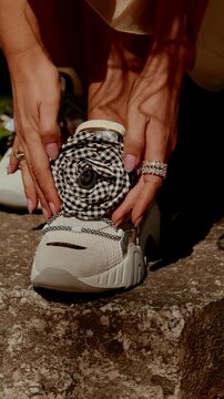 Ringed female hands adjusting decorated white sneaker with checkered fabric flower on stone