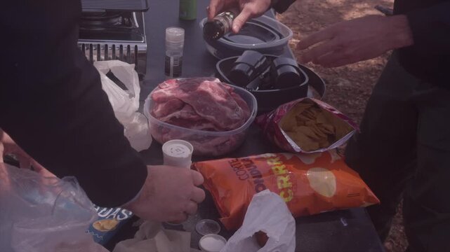 Hands sprinkling spices on raw meat in plastic container during outdoor camping picnic preparation