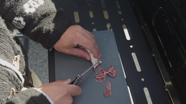 Closeup of hand slicing cured sausage on cutting board beside grill