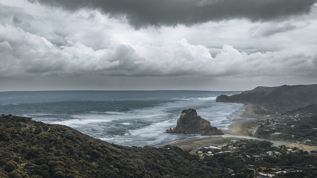Aerial view of Lion Rock and Piha Beach with crashing waves under dark moody clouds and lush green coastal hills Piha, Auckland, New Zealand.