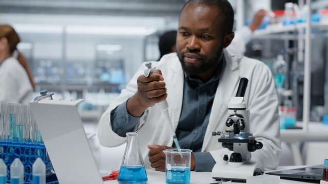 Assay development specialist pipetting reactant into blue solution for purity analysis. Man in research facility lab adding liquid agent to test tube to observe chemical reaction properties, camera B