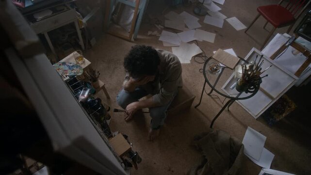 High angle view of frustrated male artist sitting among scattered sketches in messy studio, drinking whiskey and staring at canvas during creative struggle