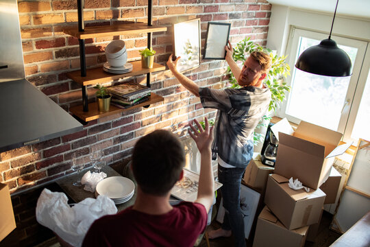 Two men hang pictures while unpacking in brick kitchen apartment