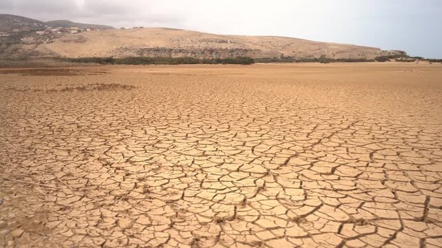 Low-angle wide shot of a vast expanse of cracked, parched earth in a severe drought landscape with a distant village. Ideal B-roll for climate change, desertification, and water scarcity projects.