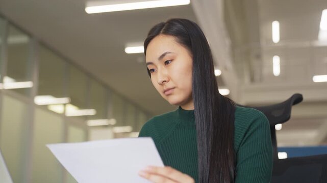 Focused Asian woman reading information on sheet of paper. Sitting in modern office with laptop open. Concentrated expression. Attention to detail. Focused on paperwork.