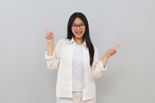 A cheerful young Asian girl looks at the camera with a smile and points to the side towards empty space, standing isolated on a white background.