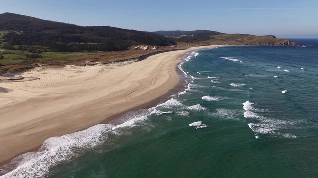 Empty sandy beach with breaking ocean waves and coastal dunes under blue sky