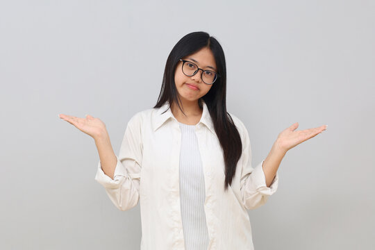 A young Asian woman with a confused and aimless expression, looking at the camera, hesitating, shrugging her shoulders and opening her palms, indicating uncertainty, stands on a white background.