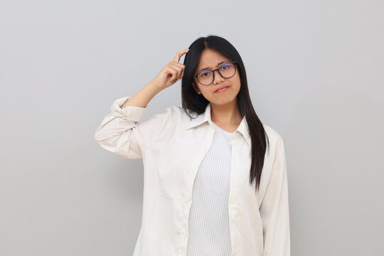 A confused looking young Asian woman wearing casual clothes, looking at the camera while scratching her head, isolated on a white background.