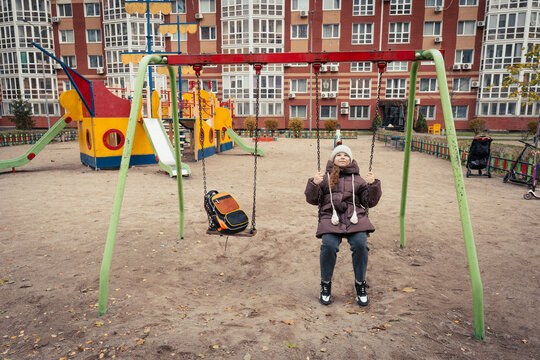 Cheerful girl wearing warm winter clothes swinging on modern playground in courtyard of apartment complex. Backpack rests on adjacent empty swing