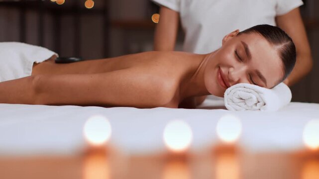 A woman with light skin relaxes on a massage table at a spa. A therapist provides a massage while candles offer soft lighting. The room has a serene and inviting feel.
