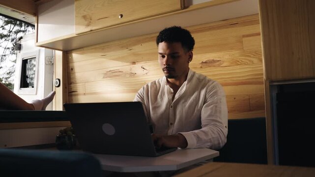 Young black man working remotely on his laptop from inside a modern camper van. His partner is relaxing on the bed in the background