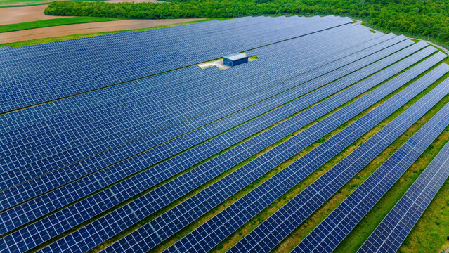 Aerial view of a large solar farm with rows of blue photovoltaic panels and a central utility building surrounded by green fields and lush forests Sabac, Vojvodina, Serbia.