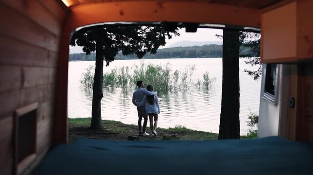 View from inside a camper van of a young couple hugging and enjoying the scenic lake view. Romantic getaway in nature for modern nomads
