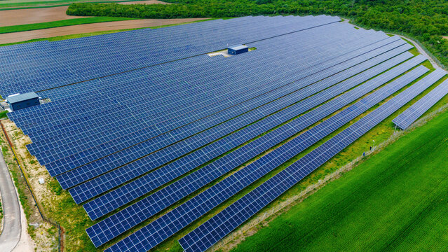 Aerial view of a large solar farm with rows of blue photovoltaic panels surrounded by lush green fields and dense forest in Sabac, Vojvodina, Serbia.