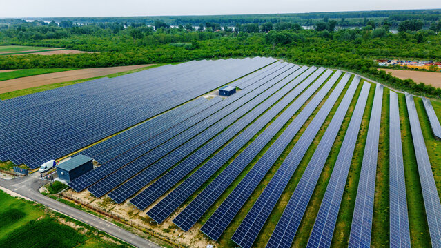 Aerial view of a large solar farm with rows of blue photovoltaic panels surrounded by lush green forests and a river in the background Sabac, Vojvodina, Serbia.