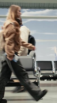 Vertical video Multiethnic men and women commuting in airport transport hub overlooked by runway control tower. Multiracial travelers walking through metal chairs inside flight departure terminal
