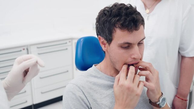 Young man sitting in a dental chair and trying on a new pair of transparent aligners. An orthodontist and her assistant are helping him