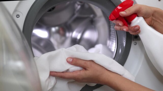 Caucasian woman prepares white laundry by spraying fabric with cleaner before placing it in a washing machine, showcasing the cleaning process in a bright laundry room