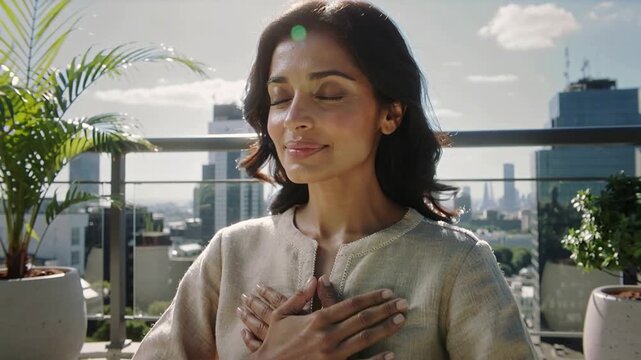 A woman sits in a quiet position on a rooftop, eyes closed and hands on her chest. The skyline of the city is visible behind her on a sunny day.