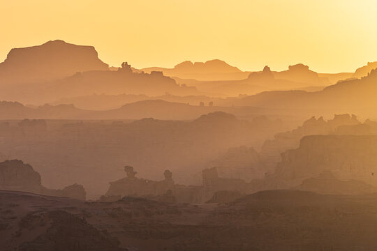 Aerial view of the Tadrart Rouge rock formations and sandstone plateaus under a golden sunset haze in Djanet, Illizi, Algeria.