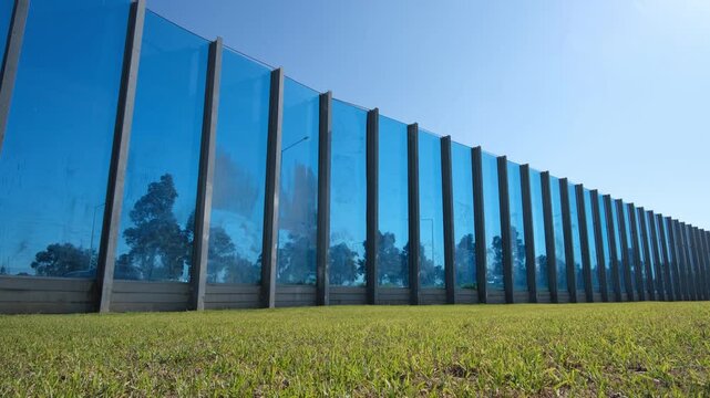 A long transparent acoustic noise barrier wall alongside a suburban highway in Melbourne, Australia with a green grass verge in foreground. Modern road infrastructure and noise mitigation technology