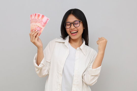 An enthusiastic young Asian woman looks happily at the camera while holding an Indonesian rupiah banknote in her hand, standing in front of a white background.
