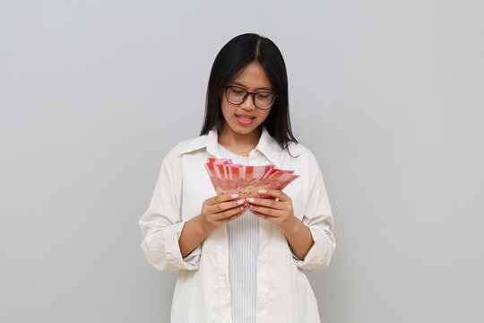 Side view photo of a young Asian woman wearing casual clothes, smiling while counting Indonesian rupiah banknotes she is holding, standing isolated on a white background.