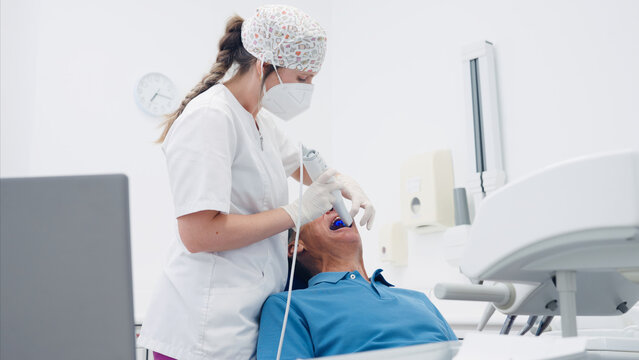 Dentist in protective mask and gloves examining male patient's teeth with advanced intraoral scanner in modern clinic