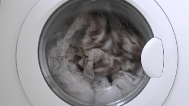Washing machine drum filled with white and patterned laundry spins rapidly, showcasing the continuous motion of clothes during the washing cycle in a bright laundry room
