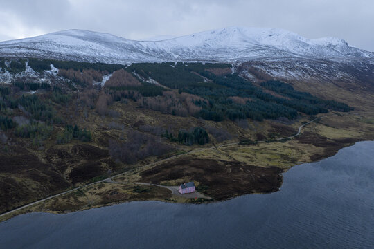 Aerial view of The Pink House on the shore of a lake with snow-capped mountains and a dense forest in Dingwall, Scotland, United Kingdom.