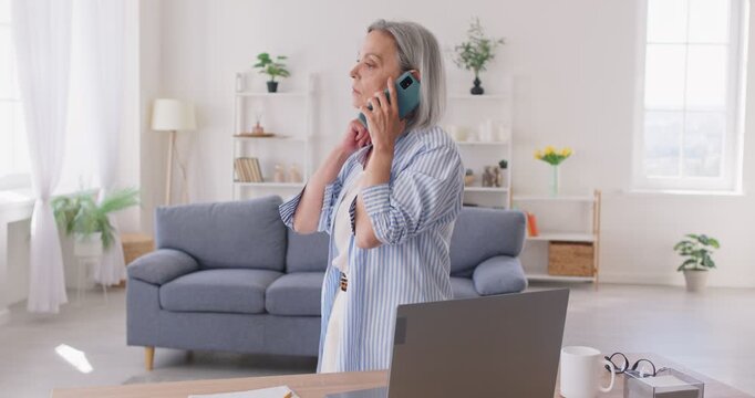 Busy mature gray haired woman working on several tasks at once at home. Senior lady holding mobile phone to her ear engaged in focused conversation surrounded by documents and laptop on her desk.