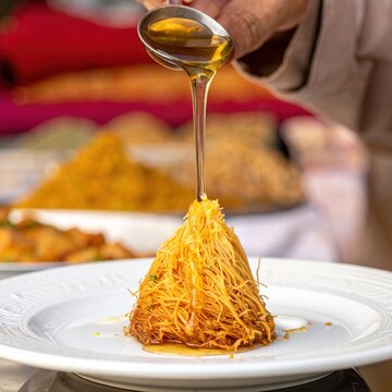 Honey syrup pouring over a shredded phyllo dough kunafa dessert, traditional middle eastern sweet pastry for celebrations and holidays