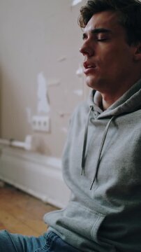 Vertical video: Sitting man in grey hoodie, blue jeans breathing after repairs in room with patches