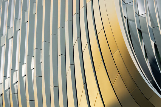 Abstract architectural detail of a modern building facade featuring curved metallic panels in gold and silver tones reflecting light in a dynamic pattern