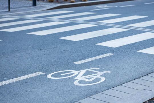 Bicycle lane marking on asphalt road with pedestrian crosswalk lines visible, urban street setting featuring gray pavement and surrounding infrastructure