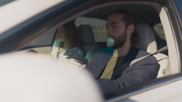 Bearded man in blazer driving car with focused expression, female passenger in striped shirt using phone beside him. Side view through open window, both wearing seat belts, sunlight shadows inside.