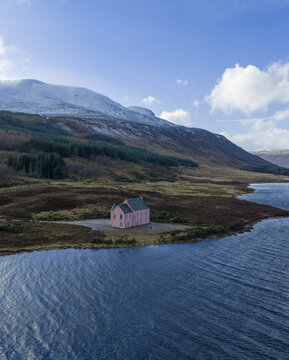 Aerial view of The Pink House on the shore of Loch Glass with snow-capped mountains and a pine forest under a blue sky in Dingwall, Scotland, United Kingdom.