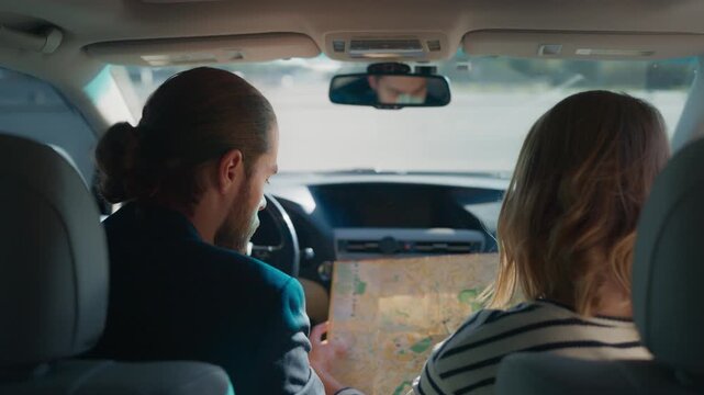 Man in blazer studying paper map while sitting in driver&rsquo;s seat, woman beside him looking at map. View from backseat inside car with bright road visible through windshield. Couple got lost. Rear.