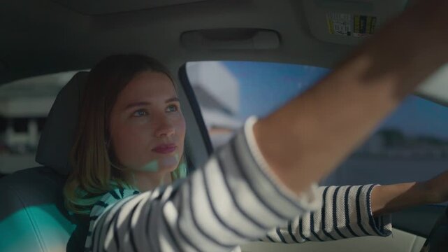 Woman in black-and-white striped sweater driving car, reaching hand to back mirror while steering. Focused gaze through windshield, short wavy hair, sunlight and blue sky visible outside vehicle.
