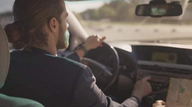Caucasian man with beard and tied-back hair driving car, adjusting dashboard controls while holding paper map. Wearing navy blazer. Steering wheel, dashboard, and road outside visible. Rear.